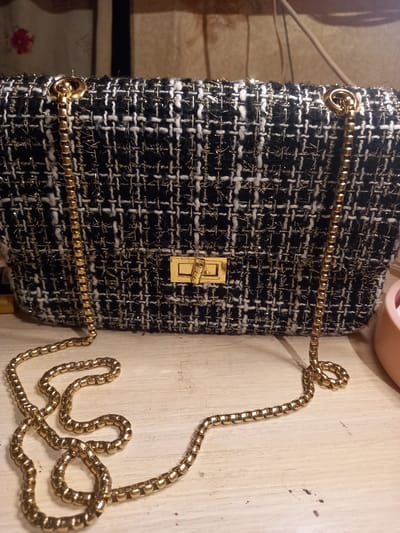 A black and white tweed handbag with gold thread accents and a gold chain strap rests on a light-colored, wooden surface. The handbag features a gold clasp closure on the front. The background consists of a textured wall hanging with a floral pattern. The scene is indoors, likely a room in Sterlibashevo, Russia. No people are visible in the image.