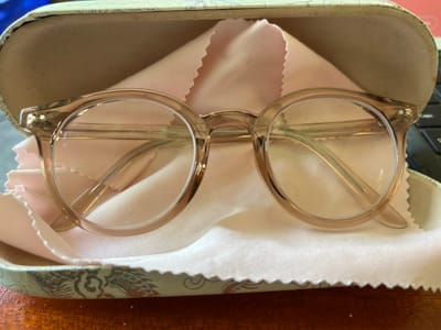 This is a medium shot, eye-level, close-up of a pair of glasses placed inside a glasses case. The glasses are round with clear lenses and a light brown frame. The frame is subtly translucent, with a slight sheen. The words "ITALY DESIGN" are visible in small, light-colored text on the inside of the right temple arm of the glasses.
The glasses are resting on a soft, pink microfiber cleaning cloth. The cloth has a slightly ruffled edge, suggesting it's designed to protect the lenses. The glasses case is partially open, revealing its tan interior. The exterior of the case is decorated with a pattern of what appears to be dragons in grey and red.
The setting appears to be indoors, with a wooden surface visible in the foreground, suggesting a desk or table. A blurred keyboard is visible in the upper right corner of the image, indicating a workspace. The lighting is neutral, suggesting it could be daytime, but the exact time is not discernible. There are no people or animals visible in the image. The overall impression is one of careful storage and maintenance of the eyeglasses. The location context of Kemloko, Indonesia, is noted, but no specific visual cues from that location are present in the image itself.