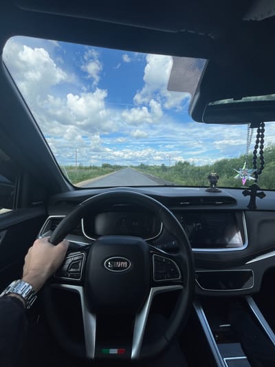 This media file is a first-person perspective shot from inside a car driving on a road, possibly in Губачево, Russia. The driver's hands are visible on the steering wheel, and they are wearing a watch. The dashboard of the car is also visible, with a digital display showing "54", which could indicate speed. There are prayer beads hanging from the rearview mirror, along with a star-shaped ornament and a cross. The car's infotainment system is displaying music information. The scene outside the car shows a road stretching into the distance, bordered by green fields and trees. The sky is bright blue with fluffy white clouds, suggesting a clear and sunny day. The image captures a sense of movement and the open road.