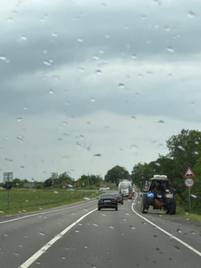The image depicts a highway scene with multiple vehicles under a cloudy sky, suggesting inclement weather, possibly rain, as indicated by water droplets on the camera lens. In the foreground, the asphalt road features a white lane divider. Several vehicles are visible: a black sedan in the left lane, a large white truck further ahead, and a blue tractor with its doors open and people inside or near it in the right lane. To the right of the tractor, a circular speed limit sign displaying "50" is posted. The roadside is lined with green grass and trees, with some vegetation and possibly distant buildings visible on the left. The general environment appears to be rural or suburban, consistent with a highway in Yaroslavskaya, Russia.
