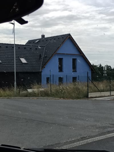 This is a medium shot, eye-level, slightly angled photograph of a bright blue house with a dark grey tiled roof. The house has a prominent gable roof with exposed wooden beams. There are two dark, rectangular windows on the upper floor of the blue section and two smaller, dark, rectangular openings on the lower floor. Attached to the left of the blue section is a darker, black house with a similar grey tiled roof and at least two dormer windows. A light grey lamppost stands in front of the black house. The property is separated from the foreground by a wire fence and overgrown dry grass. The foreground consists of an asphalt road. The sky is overcast with grey clouds. The scene is located in the village of Cvrčovice, Czechia.
