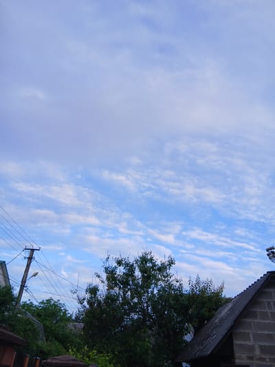 A low-angle outdoor shot captures a section of Vita-Poshtova, Ukraine, under a sky filled with wispy, horizontal clouds against a pale blue background. In the foreground, a tall, dark utility pole stands to the left, adorned with electrical wires and a single street lamp. To the right, the steeply sloped roof of a brick building is visible, its dark shingles contrasting with the grey brickwork. Lush green trees and foliage populate the midground, partially obscuring several low-rise structures with similarly dark, sloped roofs. The scene appears to be a residential or semi-rural area.