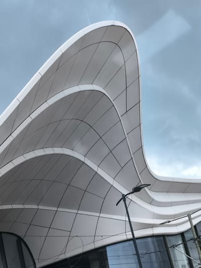 The image captures the Forum Mall, a modern architectural structure located in Leidschendam, Netherlands. The structure's design features a series of sweeping, curving white panels that form a canopy. The panels are divided into smaller, rectangular sections. The sky is overcast with a light grey-blue hue. A streetlight stands in the foreground, with its arm and light fixture silhouetted against the building. The lower portion of the building is visible with windows reflecting the surrounding environment. Electrical wires are visible near the base of the building.