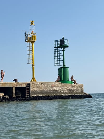 The image shows a pier with two navigational aids and several people on a clear, sunny day. On the left, a yellow navigational aid with a cage-like structure and a wind turbine on top stands tall. A woman in a bikini is on the pier to the left of the yellow navigational aid, holding a phone and seemingly taking a photo. To the right of the yellow aid, a dark, bollard-like object protrudes from the pier.
Further to the right is a green navigational aid with a solar panel. Two people are seated on the edge of the pier near the green navigational aid. One person, appearing to be a man with gray hair, is sitting with his legs extended. Another person, who appears to be a woman wearing sunglasses and a swimsuit, is sitting with her legs drawn up.
The pier is made of concrete and is weathered, with some darker material, possibly algae or marine growth, visible at the waterline. The water is a calm, blue-green color. The scene is set against a clear blue sky. This pier is likely part of the Cesenatico canal port in Cesenatico, Italy, a common location for such navigational aids and recreational activities by the sea.
