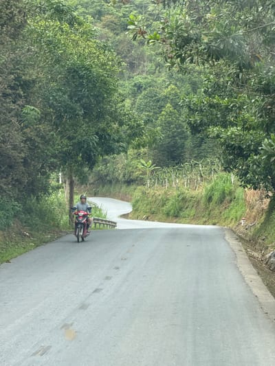 The image shows a motorcyclist on a paved road in Minh Tân Commune, Vietnam. The road curves ahead, with dense green vegetation lining both sides. The motorcyclist is wearing a helmet and riding a red and black motorcycle. The road surface shows signs of repair. The environment is lush and verdant.