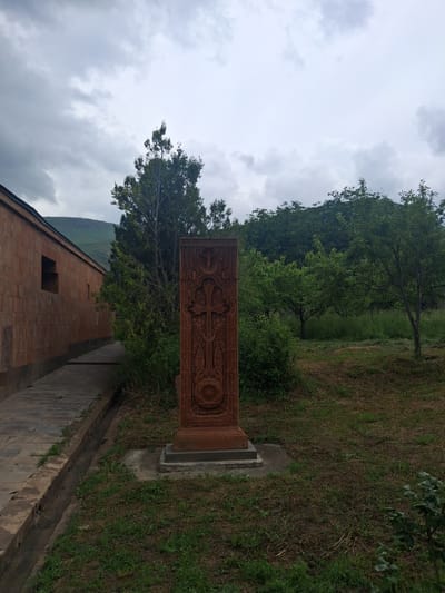 The image captures a detail of a khachkar, an Armenian carved stone cross, standing in an outdoor setting. The khachkar is made of reddish-brown stone and is intricately carved with a central cross motif and decorative patterns. It is positioned on a concrete base in a grassy area. To the left of the khachkar, a long, low building with a red-tiled roof and brown brick walls is visible. A pathway made of stone slabs runs alongside the building, bordered by a small drainage ditch. The background features lush green trees and a hilly landscape under a cloudy sky. The overall scene suggests a historical or religious site. The image was taken in the vicinity of Martiros, Armenia.