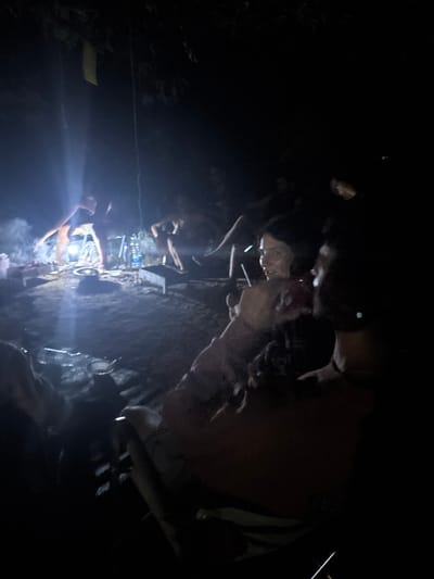 The image depicts an outdoor nighttime scene, likely a campsite or gathering, located in the Galil Elyon Regional Council, Israel. A primary light source, originating from the left mid-ground, illuminates a central area where multiple individuals are present. In the mid-ground left, one person is actively tending to a smoking charcoal grill, with visible smoke rising from it. Nearby, another individual is seated or squatting low to the ground. Several objects, including bottles and containers, are arranged on the ground around the grill. In the foreground, two individuals are partially visible, seated. One person, positioned to the right, is largely obscured. The other, a female in the mid-foreground, is illuminated and holds a beverage with a straw, looking towards the cooking area. A hand in the lower right foreground holds a thin, white object. The ground surface appears to be exposed earth or dirt. Dark tree foliage is visible in the upper sections of the frame, and a rectangular yellow fabric hangs from an unseen support in the upper left.