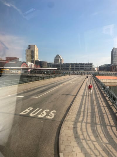 The image captures a view from a moving vehicle, likely a bus or train, looking out over a bridge in Malmö, Sweden. In the foreground, the asphalt road is marked with the word "BUSS" (Swedish for bus). To the right, a pedestrian walkway paved with light-colored bricks runs parallel to the road. A woman in a red top and beige pants is walking on this walkway, casting a long shadow. Other pedestrians and cyclists are visible in the distance on the walkway.
The backdrop features a modern cityscape. A multi-story building with a light-colored facade and balconies dominates the left side of the frame, possibly the Kronprinsen building. Adjacent to it, a building with arched windows and a red roof appears to be a train station, with the text "CYKELGARAGE" (bicycle garage) visible on a sign. Further back, a long, multi-story building with a dark facade stretches across the mid-ground, and a tall, dark skyscraper stands on the far right. Trains are visible on tracks below the pedestrian walkway. The scene is illuminated by bright sunlight, casting distinct shadows across the walkway. The sky is clear blue.