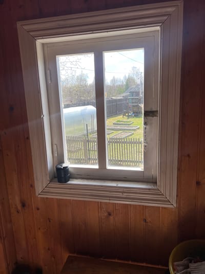 The image shows a window in a wooden building. The window is white with a decorative frame. Outside the window, there is a view of a yard with a greenhouse, a picket fence, and some garden beds. The location is specified as Чернышово, Russia. A black cylindrical object is attached to the bottom right of the window frame.