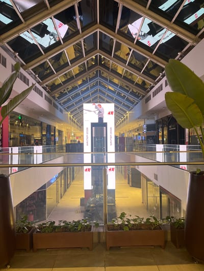 The image captures the interior of a shopping mall in Santiago de Surco, Peru. The view is directed down the center of the mall, showcasing multiple levels with stores on either side. Large advertisements for H&M with the text "WINTER 2026" are prominently displayed. The architectural design features a multi-paneled glass ceiling, allowing natural light into the space. The mall's interior includes decorative plants in large planters, adding a touch of greenery to the commercial setting. Several stores are visible along the mall's walkways, with some displaying window signage or advertising. The overall impression is a well-lit, modern commercial environment.
