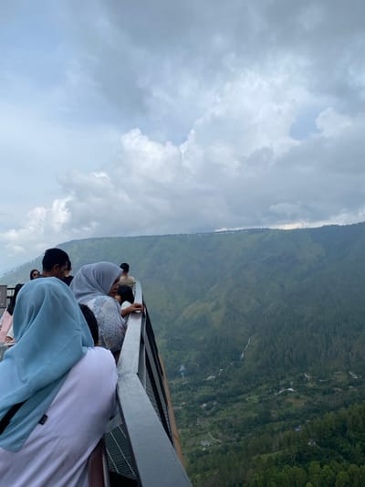 The image is a vertical, medium-angle shot taken from a vantage point overlooking a lush green valley in Turpuk Malau, Indonesia. In the foreground, a group of people stand on a viewing platform with a metal railing. A woman wearing a light blue hijab is positioned at the far left, with her back to the camera, partially obscuring a person in a white t-shirt. To her right, a woman in a grey hijab is looking down and to the right, with her hand resting on the railing. Behind them, other individuals are visible in silhouette.

The middle ground is dominated by a steep, verdant mountain slope covered in dense forest. A thin waterfall cascades down the mountainside. The valley floor is dotted with scattered houses and a winding road. The background features the undulating peaks of more mountains, shrouded in a misty haze under a heavy, overcast sky filled with grey and white clouds. The overall atmosphere is serene and natural, with a hint of human presence on the viewing platform.