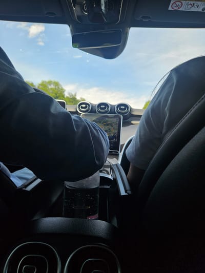 This media file shows the interior of a car, looking out through the windshield. The scene is shot from a low angle, and the perspective is rotated, making it appear as if the car is on its side. The primary subject is the interior of the car, with a focus on the dashboard and the view outside.
In the foreground, a person's legs are visible, clad in dark pants. They are sitting in the passenger seat, with their knees bent. Part of a person wearing a white shirt is visible in the background, seemingly the driver.
On the dashboard, a prominent touchscreen display is visible, showing a map with navigation details. There are also several circular air vents, a common feature in car interiors. A dark-colored object, possibly a water bottle or a container, is placed in a cup holder next to the gear shift.
Through the windshield, a bright blue sky with a few scattered white clouds is visible. Green trees can be seen outside, suggesting a location with natural surroundings. The lighting indicates it is daytime, likely late morning or afternoon, with clear weather.
The orientation of the image suggests it was taken in France, as indicated by the location context of Châtenay-Malabry. There are no discernible emotions conveyed, as the focus is on the setting and objects. A safety warning sticker is partially visible on the sun visor in the top left of the image, but the text is too blurry to be read.