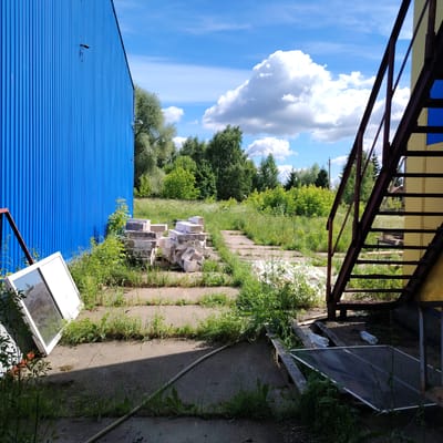 The image shows an outdoor scene in the village of Rakhmanovo, Russia. In the foreground, there is a concrete pathway overgrown with grass. To the left, a large blue corrugated metal building dominates the frame. In front of the building, a white window frame with glass lies on the ground. A pile of white building materials, possibly bricks or blocks, is situated on the pathway. To the right, a rusty metal staircase with yellow siding leads upwards. The background features lush green trees and foliage under a bright blue sky with fluffy white clouds. The overall impression is one of an industrial or construction site that is partially overgrown with vegetation.