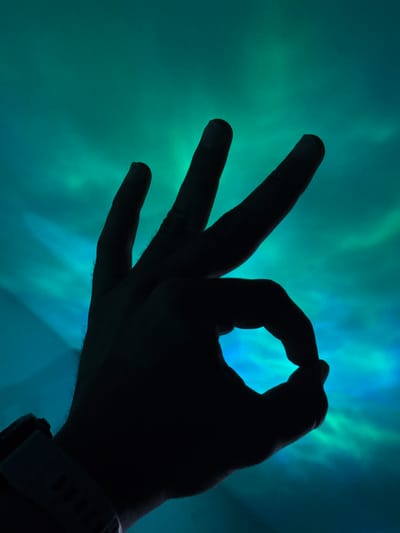 A silhouetted hand forms an "OK" gesture against a vibrant, swirling backdrop of blue and green light, suggestive of the aurora borealis. The hand, with fingers extended and thumb and index finger forming a circle, is dark and lacks discernible features. A watchband is visible on the wrist. The background is characterized by ethereal streaks and clouds of luminous color. No specific location in Santa Marta, Colombia, can be identified from the image.