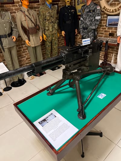 A collection of mannequins dressed in various military uniforms stands against a brick wall. A machine gun is mounted on a green-topped display table. The table includes explanatory text and an image depicting a similar gun. The angle of the photo provides a view down the barrel of the gun, with the mannequins in the background appearing small.