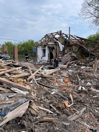 The image shows the aftermath of an explosion in Bilopillia, Ukraine, at the coordinates 51.139 latitude and 34.286 longitude. A single-story residential building has been heavily damaged and is partially collapsed. The debris of the structure is scattered throughout the yard, and there are signs of fire damage. A person in a black and yellow uniform, likely a firefighter or rescue worker, is standing in the yard near the rubble. There is also a person in a black shirt standing on top of the building, surveying the damage. The yard is covered in debris, dirt, and mud, and the sky is overcast. The scene is chaotic and destructive, highlighting the impact of the explosion.