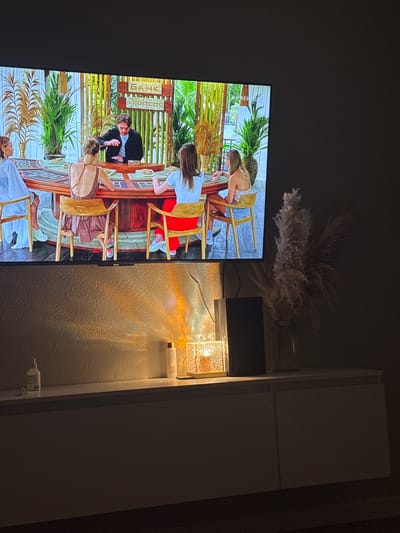 A widescreen television displays a scene from what appears to be a game show or reality television program. Five people are seated around a large, circular table with a red surface. A man in a dark suit jacket stands behind the table, interacting with the participants. On the table are what look like circular tokens or chips. Behind the table, a decorative backdrop features tall, slender plants and a sign with Cyrillic lettering that reads "БАНК" (BANK) and numbers. The television is mounted on a wall above a white media console. On the console, several objects are placed, including a clear glass vase with dried decorative grass, a dark rectangular object (possibly a speaker or soundbar), a glass lamp with a textured shade emitting a warm light, and two bottles. The room is dimly lit, with the primary light source coming from the lamp and the television screen, casting shadows and reflections on the textured wall behind the console. The setting appears to be a domestic environment, possibly a living room, with no discernible urban landscape from the city of Kuyuki, Russia, visible within the frame.