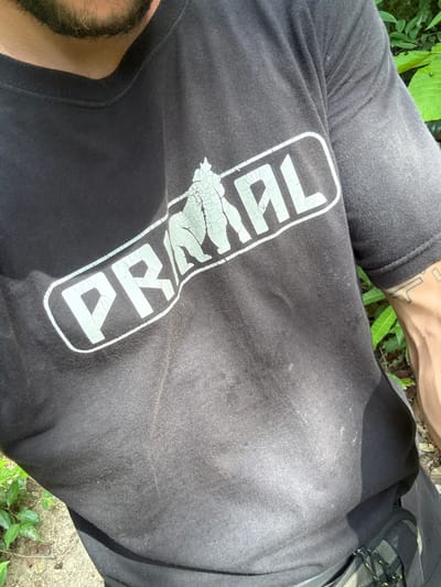 An overhead shot of a person wearing a black t-shirt with a white logo that reads "PRΛNΛ" with cracked print. The person is also wearing grey jeans. A small portion of a boot is visible on the right side of the frame. A bearded chin is visible on the bottom left corner. Tattooed forearm on upper left. Vegetation and concrete surface are also visible. The image was taken in Moo 7, Thailand.