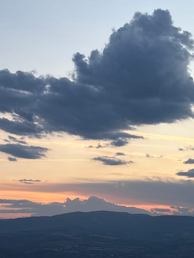 The image captures a dramatic twilight sky, likely a sunset or sunrise, over a mountainous landscape in Elenovo, Bulgaria. The scene is dominated by a vast and dense mass of dark grey clouds occupying a significant portion of the sky, suggesting an approaching storm or heavy weather system.
To the right, the sky presents a striking contrast with bands of warm, golden-yellow light transitioning into soft orange and pink hues along the horizon. Scattered, lighter clouds are visible in this brighter section, some catching the sun's glow while others appear as darker wisps. The interplay of light and shadow among these clouds creates a dynamic and textured overhead display.
Below the illuminated horizon, a dark silhouette of distant mountains or hills stretches across the lower part of the frame, appearing indistinct against the vibrant sky. The overall impression is one of natural grandeur and the powerful interaction between weather elements and atmospheric light. There are no visible people, objects, activities, or text within the image. The scene vividly portrays the dramatic beauty of nature during twilight, with its stark contrasts and evolving colors.