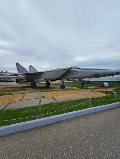 The image shows a MiG-25 fighter jet on display at the Vadim Zadorozhny Technology Museum in Arkhangel’skoye, Russia. The aircraft is painted in a light gray color scheme with the number "47" painted in blue on the side. The jet is positioned on a concrete pad next to a grassy area and a paved road. A low concrete barrier runs along the edge of the paved road, and chain link fencing provides a boundary to the display area. The sky is overcast.