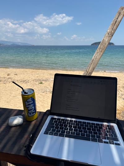 This media file captures a serene outdoor work scene by the sea. The primary subjects are a laptop, a can of Fanta, and a wireless mouse, all placed on a wooden surface. In the background, a tranquil beach transitions into the clear turquoise waters of the sea, with a hazy coastline and islands visible in the distance under a bright blue sky dotted with white clouds. A weathered wooden post extends from the left of the frame, suggesting a rustic beachside setting.
The scene evokes a sense of peaceful productivity, with the gentle lapping of waves and the warm sunshine creating a relaxed atmosphere. The laptop is open, displaying a document with dialogue snippets, indicating that someone is engaged in writing or editing. The Fanta can, with its distinctive yellow and blue branding, and a black straw, suggests a refreshing break. The overall impression is one of a person enjoying a work-from-paradise scenario.
The time of day appears to be late morning or early afternoon, given the bright sunlight casting distinct shadows. The weather is clear and sunny, ideal for outdoor activities. The location cues, such as the sandy beach and the characteristic Greek island silhouette, point towards a Mediterranean coastal setting, specifically Νέα Ηρακλείτσα, Greece. There are no people visible in the frame, focusing the attention entirely on the workspace and the natural beauty surrounding it. The visible text on the Fanta can and within the laptop document provides specific details about the brand and the content being worked on.