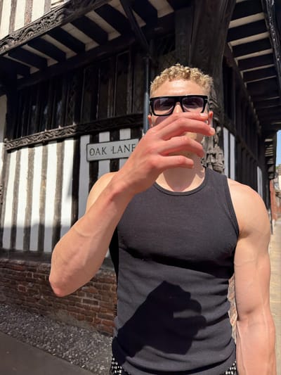 The image shows a person in a black tank top with a tattoo on the left arm, obscuring their face with their hand. The person is wearing black-framed glasses and has blonde curly hair. The location appears to be Ipswich, United Kingdom, based on the Oak Lane street sign and the architecture visible in the background, which includes a Tudor building with black and white timber framing. The setting is outdoors.