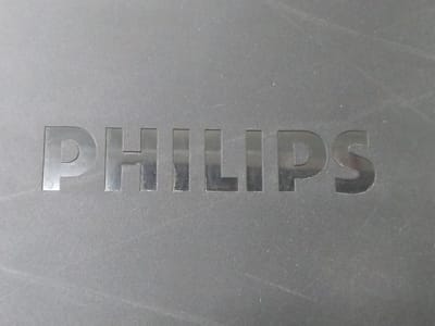 The image is a close-up shot of the word "PHILIPS" embossed on a dark gray, textured surface. The letters are in a metallic silver color and appear to be recessed into the surface. The lighting casts subtle shadows, highlighting the embossed nature of the lettering. The background is uniformly dark gray with some faint scratches and dust specks visible, suggesting the object is likely a piece of electronics or an appliance. There are no people or any other distinguishing environmental features visible in the frame.