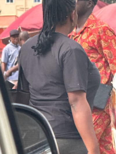 The image shows a street scene in Orji, Nigeria. In the foreground, a person wearing a dark t-shirt is partially visible from behind. Behind them, a person in a red and gold patterned shirt is also visible. Further in the background, another person wearing a striped shirt and a hat is seen. A red umbrella is partially visible in the upper portion of the image. The environment appears to be an outdoor setting, possibly a market or street vendor area, with buildings and other people in the background.