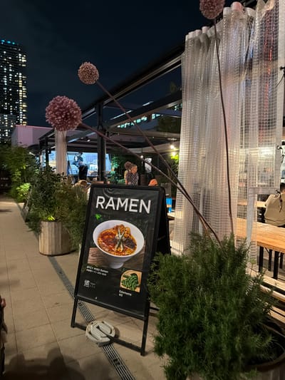 A black A-frame sign displaying "RAMEN" with an image of ramen and "SPICY MISO Ramen" text is positioned on a tiled walkway. To the left of the sign, a tall building with many illuminated windows is visible against a dark night sky. Further left, lush green plants in wooden planters line the walkway. Behind the sign and plants, a covered outdoor seating area is discernible with tables and people dining. A person wearing a blue shirt and an orange hat is partially visible in the background. To the right of the sign, a large, leafy green plant is in a dark pot. Translucent white curtains hang from a structure behind the plant. A wooden bench with people sitting at it is also visible in the background to the right.