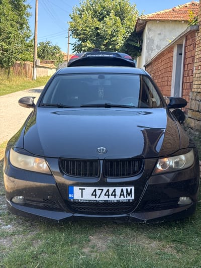 A black BMW sedan is parked on a grassy area next to a dirt road. The car faces the camera. The license plate reads "T 4744 AM" and has "BG" on the left, indicating Bulgaria. The car's headlights are on. Behind the BMW, another car, a dark color, is partially visible. The background shows a brick building with a tiled roof, trees, and a wooden fence. The environment appears to be rural. The image was taken in the city of Konop, Bulgaria.