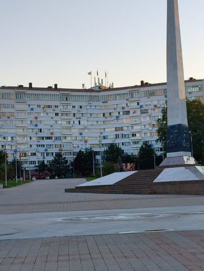 The image depicts a large, curved, multi-story apartment building with numerous windows, balconies, and visible air conditioning units. In the foreground, a tall, gray obelisk monument stands prominently, adorned with a dark, sculpted relief at its base. The monument is surrounded by a paved plaza with tiered steps and angled platforms. Trees and some flowering bushes are visible in the mid-ground, between the monument and the building. The sky above is clear and light. The scene appears to be an urban public space, likely a monument or memorial square within the city of Agoy, Russia, given the prompt's context. There are no people visible in the image.