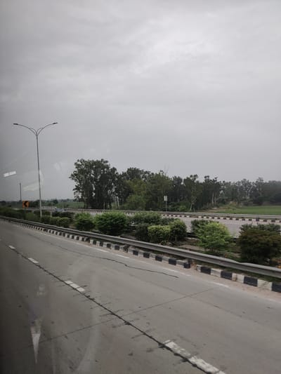 The image shows a road with multiple lanes, bordered by a metal guardrail with black and white markings. Beyond the guardrail, there is a row of green bushes and trees. Further in the background, a dense line of tall trees is visible, with some greenery and fields interspersed. The sky is overcast and grey, suggesting a cloudy day. A tall street lamp with two arms is present on the left side of the road. There is a yellow directional sign with a black arrow on the left side of the road. The road itself is paved and appears to be in use, with white lane markings. The overall scene suggests a highway or a major road in a semi-urban or rural area, possibly in India given the vegetation and style of the street lamp.