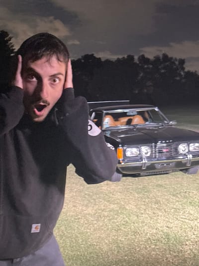 A startled man stands with hands over his ears, mouth agape, with wide eyes gazing at the camera. He is wearing a black hooded sweatshirt with a Carhartt logo. Behind him is a vintage black car with tan leather seats parked on a grassy area. The sky is overcast and the photo appears to be taken outdoors at dusk or night. Trees are visible in the distance.