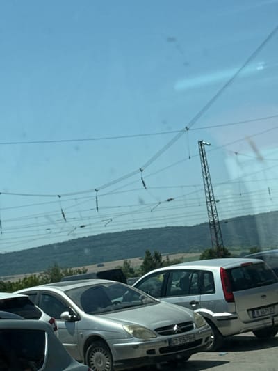 The image shows a view from a vehicle looking out at a parking lot filled with cars. In the foreground, several cars are visible, including a silver Citroën C4 with license plate CT 9557 BH. To the right of the Citroën is a silver Ford Focus. Behind these cars, a tall, metal lattice structure, likely a pylon for overhead power lines, is visible against a clear blue sky. In the background, a forested hillside slopes down towards the parking lot. The scene appears to be a public parking area in Dalgopol, Bulgaria, indicated by the Bulgarian license plates on the vehicles. The lighting suggests it is daytime.