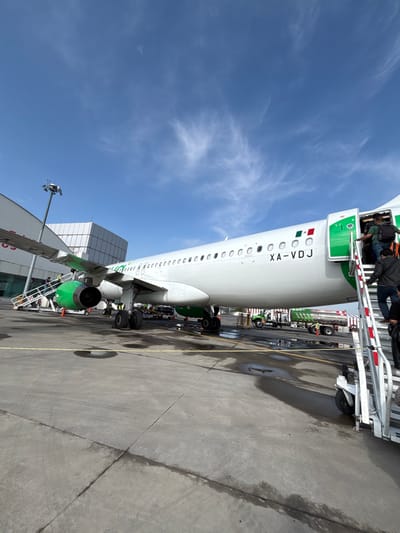 An outdoor, daytime, low-angle full shot captures an airplane at an airport in San Miguel Totoltepec, Mexico. The white airplane, with green accents on its wingtips and engines, is parked on a concrete tarmac. The aircraft's registration number, "XA-VDJ," is visible on its fuselage, along with the Mexican flag. Passengers are boarding the plane via a metal staircase attached to the open door at the rear of the aircraft. Several airport service vehicles, including a fuel truck, are parked near the airplane. The sky above is a clear blue with scattered wispy clouds.