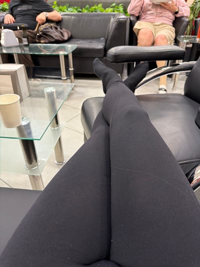 In a waiting area, possibly a salon, a person is seated with their legs crossed. They are wearing black leggings and dark socks. In the background, another person is seated on a black leather couch, holding a brown purse. A glass-top table with metal legs sits between the two individuals. The environment appears to be indoors, with a potted plant visible in the background. The scene does not provide any specific clues to pinpoint its location within Khrabrovo, Russia.