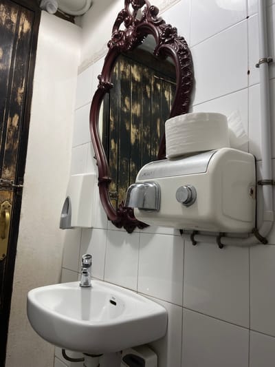 The image depicts a white tiled bathroom in Agüimes, Spain (latitude: 27.907, longitude: -15.447). A white porcelain sink with a chrome faucet is positioned against the wall. Above the sink is a white electric hand dryer mounted to the wall. A roll of paper towels is visible on top of the hand dryer. A large ornate wooden framed mirror hangs on the wall above the dryer and sink, reflecting a wooden door. The bathroom is clean and well-lit. The scene is devoid of people.  The image is taken from a slightly low angle.  There are no other objects or actions in the image.