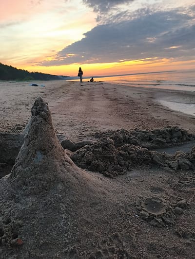 This image captures a serene beach scene in Apšuciems, Latvia, during either sunrise or sunset. The foreground is dominated by a meticulously crafted sandcastle, its conical structure textured and detailed. Scattered around it are piles of damp sand, suggesting ongoing construction or recent play. In the mid-ground, the sandy expanse stretches towards the water, marked by the footprints and scattered debris of beachgoers. A lone figure stands near the water's edge, silhouetted against the soft glow of the sky, while another individual is seated further down the beach. The Baltic Sea, calm and reflecting the warm hues of the sky, meets the shore gently. The horizon is lined with a dark, dense forest, contrasting with the open sky above, which is a canvas of soft oranges, yellows, and blues, streaked with dramatic, silhouetted clouds. The overall atmosphere is one of peaceful tranquility and the simple joys of a day at the beach.