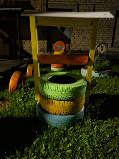 The image depicts an outdoor scene at night, likely in a residential area. In the foreground, a structure resembling a well or a playhouse is constructed from painted tires. Three tires are stacked vertically, painted green, yellow, and blue from top to bottom. A yellow wooden frame supports a white roof over the tires. To the left, two orange tires are placed on the grass. In the background, a brick building is visible, with a dark doorway. A sign with a cartoonish face, possibly a Mickey Mouse character, is attached to the yellow frame. The ground is covered in grass, which is illuminated by artificial light, creating shadows. The overall impression is of a DIY or repurposed playground feature.