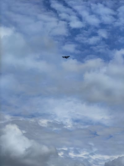A small, dark-colored biplane is captured in flight against a sky filled with a mix of white and gray clouds. The plane is positioned in the upper center portion of the image. The sky is a pale blue, with varying shades of clouds indicating potential weather changes. The image does not show any specific landmarks or indications of its location within Яглевіцкі сельскі Савет, Belarus.