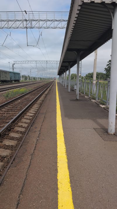A low-angle, long shot captures a train station platform in Elban, Russia, under a cloudy sky. The platform itself is asphalt with a bright yellow line marking the edge, and a covered waiting area supported by grey pillars runs along the right side of the frame. To the left, railroad tracks stretch into the distance, with a train of green cargo cars visible on one set of tracks. Overhead, a complex network of power lines and supporting structures for the electric railway crisscrosses the sky. The environment appears to be an operational, albeit somewhat weathered, rural train station.