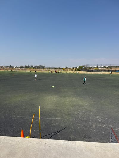 An outdoor artificial turf sports field in La Venta, Mexico, is depicted under a clear blue sky. In the immediate foreground, a concrete ledge partially obstructs the view of an orange cone and two vertical yellow training poles. The dark green and grey artificial turf extends across the midground and background. One individual, dressed in a white long-sleeved shirt and white shorts, stands in the left-center midground, facing forward. To their right, another individual, wearing a dark jacket, light trousers, and a cap, walks from right to left across the field. A crumpled light green object rests on the turf between these two individuals. In the distant background, multiple groups of people are visible, some gathered near white goalposts, indicating a soccer practice or event. Trees, distant buildings, and a tall mesh fence line the far horizon on the right. Shadows are cast by the foreground objects, suggesting bright overhead sunlight.