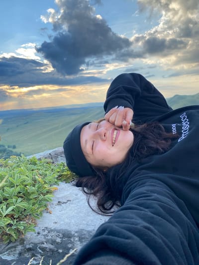 A young woman lies on her back on a rocky surface with green vegetation growing on it, facing the camera. She is wearing a black beanie and a black hooded sweatshirt. Her eyes are closed, and she has a smile on her face, with her right hand covering her forehead. She has a nose ring. The background consists of a vast, rolling green landscape under a cloudy sky with hints of sunset light filtering through. The scene appears to be in a natural, elevated location, possibly a mountain or high plateau. The location is identified as Abzanovo, Russia.