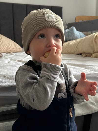 An indoor, chest-up shot depicts a young child in the city of Gribovo, Russia. The child, wearing a light brown ribbed knit beanie with a white rectangular patch displaying a rainbow logo, a grey long-sleeved sweater with striped cuffs, and dark blue overalls with dark shoulder straps, is holding a small, light-colored, round food item, resembling a cookie or biscuit, to their mouth with their right hand. Their left hand is open with fingers slightly splayed, positioned to the left of their torso. The child's blue eyes are directed upwards and to the left. The background features a bed with light-colored bedding, including a white textured cover and beige pillows, situated against a dark grey upholstered headboard with vertical paneling. Partially visible furniture, including a wooden crib or bed frame with a dark grey cushion and a light blue object, is present in the deeper background to the right.