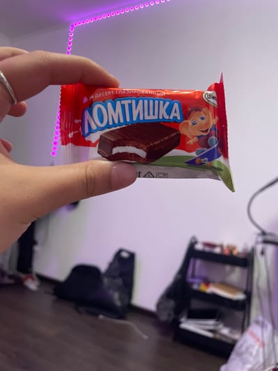 The image shows a hand holding a wrapper for "Lomtiyshka" glazed dessert. The wrapper is red and features a cartoon character of a bear on a skateboard. The dessert itself, a chocolate-covered biscuit with a white cream filling, is visible through the clear plastic part of the wrapper. The background is out of focus, but appears to be an indoor setting with a shelf unit and some dark bags on the floor. There are also pink LED lights visible on the wall. The location of Borisova Griva Massiv, Russia is not depicted in this image.