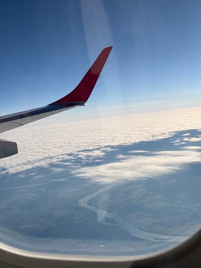 The image shows an airplane wing, seen through a window, over a landscape. The wing is red on the tip and gray below. A river is seen in the background, with what appears to be some land masses near it. The image may have been taken near Schwechat, Austria, based on the possible presence of the Danube River. Above the river there seems to be a layer of clouds.