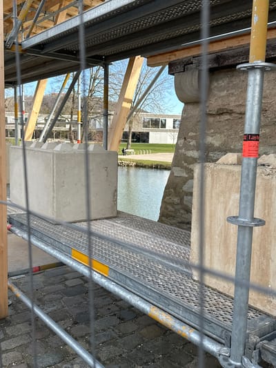 The image displays an active construction or renovation site in Weißenburg in Bayern, Germany, on a clear, sunny day. The foreground is occupied by a metallic mesh fence, through which the scene is viewed. Behind the fence, a cobblestone surface is visible. A multi-level metal scaffolding system, including vertical poles (one bearing a "Layher" sticker) and perforated metal platforms with yellow reflective strips, is prominent. On a central platform, a large, light grey rectangular concrete block with conical protrusions on its upper surface rests. The scaffolding system appears to enclose or support a weathered stone structure, possibly a historical bridge abutment or building foundation, partially visible on the right. In the midground, a calm body of water, likely a river or canal, reflects the bright blue sky. On the distant bank, a green grassy area features a bicycle rack, a white bench, and two distant human figures—one seated, one standing. Further back, modern buildings with large windows are visible amidst bare trees, with distant hills under a clear blue sky.