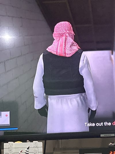 The image shows the back of a person wearing a white thobe and a black vest. They are also wearing a red and white checkered keffiyeh. The person appears to be standing in an indoor setting with a brick wall in the background. There is text on the screen that reads "Take out the d". The image quality is poor, suggesting it is a photograph of a screen. The image does not contain any information about the city of Boboshevo, Bulgaria.