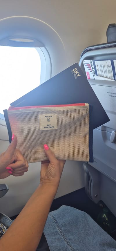 A person's hands are holding an open, beige mesh pouch with a pink zipper and a small white label. The pouch has "APIVITA BEE SUN SAFE" written on the label. Underneath the pouch is a dark blue folder with the "SKY EXPRESS" logo and "skyexpress.com" visible. The scene appears to be inside an airplane cabin, with a window visible on the left and the overhead compartment of an airplane seat on the top. A person wearing a denim jacket is partially visible on the right. The lighting suggests it is daytime, likely during a flight. The text "320neo" is visible on a card above the folder. Other text on the card is in Greek and English, with instructions related to electronic devices on an airplane. The overall impression is of someone holding travel amenities or personal items during a flight.