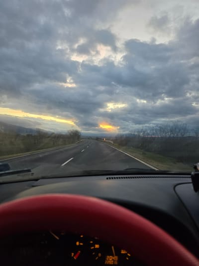 The image shows a view from inside a car driving on a road. The sky is predominantly cloudy with patches of sunlight breaking through. The road ahead stretches into the distance. The dashboard and part of the steering wheel are visible. Based on context clues, the car is located near Osetenovo, Bulgaria, implied from the metadata. There is no presence of people or interactions in the frame.